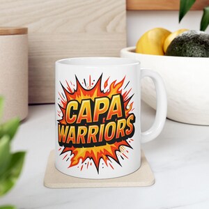 May include: White ceramic mug with the text "CAPA WARRIORS" in a comic book style design. The text is in orange and yellow, outlined in black, set against a red and yellow burst graphic. The mug has a white handle and sits on a square coaster.