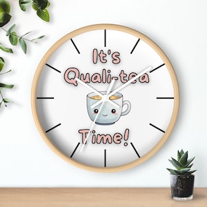 May include: White round clock with black hands and black markings. The clock has a wooden frame and a cute cartoon teacup with a smiling face. The text on the clock reads "It's Quali-tea Time!"