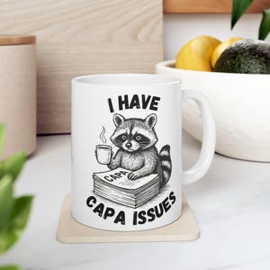 Puede incluir: Taza de cerámica blanca con una ilustración en blanco y negro de un mapache sosteniendo una taza de café y sentado sobre una pila de papeles. La taza tiene el texto "I HAVE CAPA ISSUES" en negro.