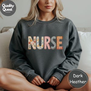 May include: Dark grey sweatshirt featuring the word "NURSE" in a patchwork design. The letters are decorated with floral, plaid, and geometric patterns. The sweatshirt is a crew neck style.