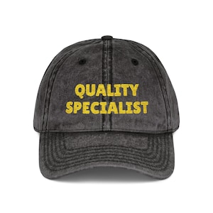 May include: A faded black baseball cap featuring the text "QUALITY SPECIALIST" in yellow. The cap has a curved peak and a button at the crown, suitable for casual wear.