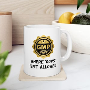 Peut inclure: Une tasse en céramique blanche avec un sceau or et noir qui dit "GMP Certified" et le texte "Where 'oops' isn't allowed".