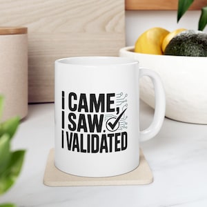 May include: White ceramic mug with the text "I CAME, I SAW, I VALIDATED" in black, with a checkmark and circuit board design. The mug sits on a beige coaster.