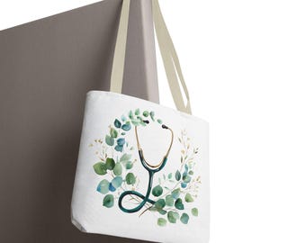 May include: White tote bag featuring a watercolor design of a stethoscope surrounded by green eucalyptus leaves and gold accents. The bag has beige straps and is displayed against a neutral background. A medical-themed accessory.