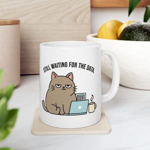 Puede incluir: Taza de cerámica blanca con un diseño de gato de dibujos animados, un ordenador portátil, una calculadora y una taza de café. El texto "STILL WAITING FOR THE DATA" está impreso encima del gato. La taza está sobre un posavasos beige.