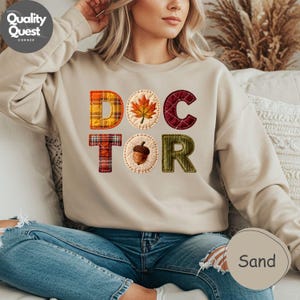 May include: A sand-coloured sweatshirt displaying the word "DOCTOR" in a patchwork design. The "O" is a maple leaf, and the "O" has an acorn. The sweatshirt is a comfortable, casual style.