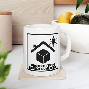 May include: White ceramic mug with a black graphic of a house, sun, and cube, with the text "PROTECT FROM DIRECT SUNLIGHT". The mug is on a beige coaster, with a blurred background of a wooden surface and a bowl of fruit.