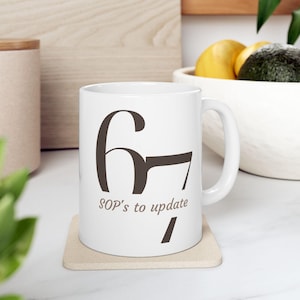 Może przedstawiać: Biały ceramiczny kubek z brązową grafiką liczby 67 i napisem "SOP's to update". Kubek stoi na beżowej podstawce. Uchwyt jest widoczny po prawej stronie kubka.