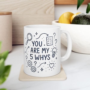 Peut inclure: Mug en céramique blanche avec l'inscription "YOU ARE MY 5 WHYS" en noir, accompagnée d'illustrations de loupe, d'ampoule, d'engrenages et de points d'interrogation. Le mug est posé sur un dessous de verre beige.