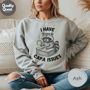 May include: Pale grey sweatshirt with a raccoon graphic holding a coffee cup and sitting on books. The text "I HAVE CAPA ISSUES" is printed above and below the raccoon illustration. A relaxed-fit, comfortable sweatshirt.