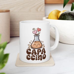 Könnte beinhalten: Weiße Keramik-Tasse mit einem Cartoon-Becher-Design mit Herzen und dem Wort "CAPACCINO" in braun. Die Tasse steht auf einem beigen Untersetzer.