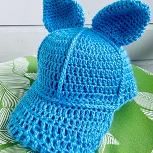 May include: A bright blue, crocheted baseball cap with two large, upright ears. The cap features a curved brim and is made of a textured, open-weave fabric. The hat is designed to resemble an animal.