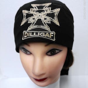 May include: Black head wrap featuring a silver and black cross design with a skull in the center. The head wrap has the text "DILLIGAF" embroidered on the front. The head wrap is worn by a mannequin.