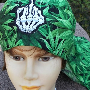 May include: A green head wrap featuring a pattern of cannabis leaves. A white and black patch of a skeleton hand giving the middle finger is centered on the front. The head wrap is made of a soft fabric.