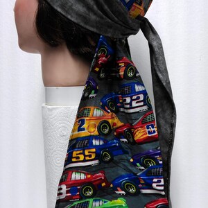 May include: A gray head wrap with a racing car pattern. The fabric features colorful race cars with numbers like 2, 22, and 55. The head wrap is tied at the back with long, gray fabric ties.