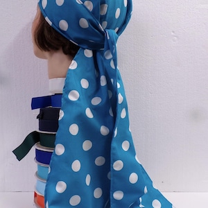 May include: A blue headscarf with white polka dots is tied at the back. The scarf is draped over a mannequin head. A stack of colorful ribbons is visible in the background.