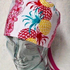 May include: White surgical scrub cap with a vibrant tropical print. The pattern features red and yellow pineapples with teal leaves, and pink flowers. The cap has a pink tie in the back.