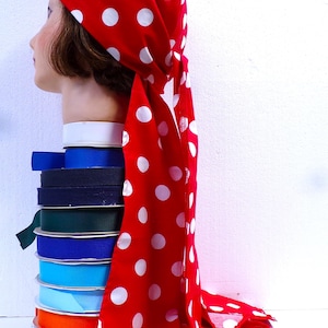 May include: A red headscarf with white polka dots is draped over a mannequin head. A stack of colorful ribbons in various shades of blue, green, and orange sits below the head. The headscarf's fabric is flowing.