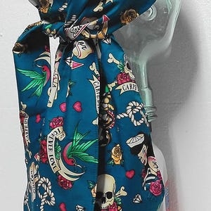 May include: A teal head wrap featuring a tattoo-style print with skulls, red roses, playing cards, swallows, and ropes. The fabric includes text banners reading Carpe Diem and Forever Love, with small red hearts scattered across the patterned surface.