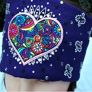 May include: A dark blue headband featuring a large, embroidered heart filled with colorful floral designs. The heart is outlined in pink and adorned with sparkling rhinestones. Additional rhinestones and white paisley patterns decorate the headband.