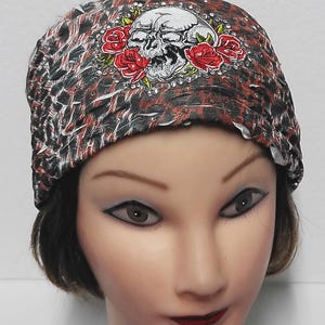 May include: A black, red, and white patterned headband featuring a skull and red rose design. The skull is white with black detailing, and the roses are a vibrant red. The headband is worn on a mannequin head.