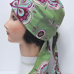 May include: A green head covering with a paisley pattern in shades of pink, teal, and white. The head covering is tied at the back with long, flowing ties. The fabric has a smooth texture.