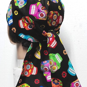 May include: Black head covering featuring a colorful sugar skull pattern. The skulls are in various colors including pink, blue, green, and red, with floral and decorative accents. The head covering is tied at the back.