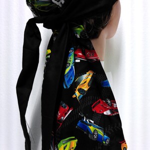 May include: A black head wrap with a racing car print. The head wrap features a variety of colorful race cars on a black background, with a section of white cars on a black background. The wrap has long black ties.