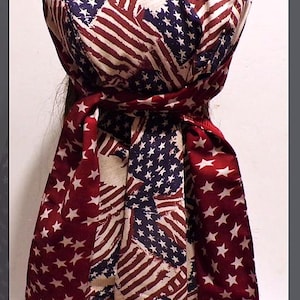 May include: A head wrap featuring a patriotic American flag design. The fabric displays the stars and stripes in red, white, and blue. The wrap is tied at the back, with long, flowing fabric.