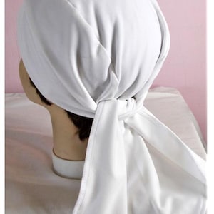 May include: A white head covering, likely a scarf or bonnet, is tied at the nape of the neck. The fabric is smooth and appears to be a soft material. The head covering is draped over a mannequin head.