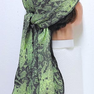 May include: A green and black snake print durag. The durag is tied at the back and has long, flowing tails. The fabric has a textured appearance. The durag is designed to be worn on the head.