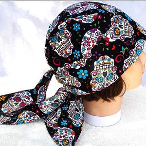 May include: A black head wrap featuring a colorful sugar skull pattern. The design includes white, blue, red, and yellow floral and skull motifs. The head wrap is tied at the back with long, patterned tails.