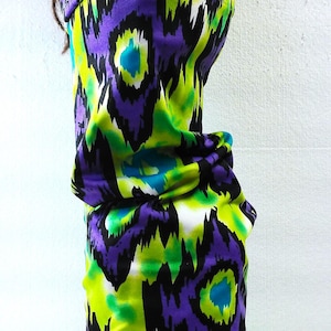 May include: A tubular neck gaiter featuring a vibrant, abstract ikat-inspired pattern in shades of lime green, deep purple, and bright turquoise. The fabric is lightweight and stretchy, designed to cover the neck and lower face area completely.