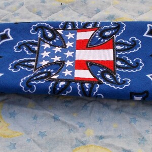 May include: A blue bandana with a white paisley pattern and a central patch featuring the American flag and a Maltese cross design. The bandana is folded, showcasing the design.