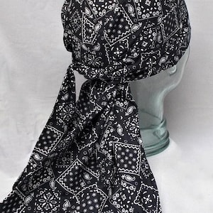 May include: Black bandana-style head covering with a white paisley pattern. The head wrap is tied at the back, with long fabric tails hanging down. The fabric is soft and appears to be cotton. The head covering is designed to fit over the head.