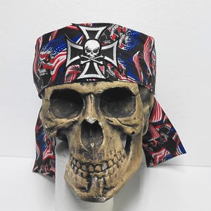 May include: A black bandana with a white skull and crossbones emblem, a white cross, and red, white, and blue American flag patterns. The bandana is draped over a skull-shaped decorative object.