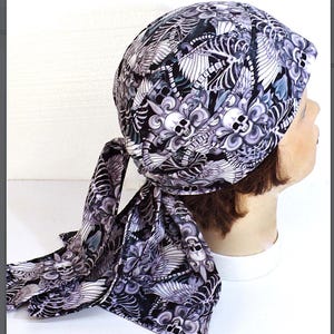 May include: A black, white, and gray bandana featuring a repeating pattern of skulls, wings, and floral elements. The bandana is tied at the back, with long tails hanging down. The head covering is made of a soft fabric.