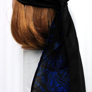 May include: Black head wrap with a blue paisley design. The head wrap is tied at the back and covers the head. The paisley pattern is a repeating design of curved shapes and teardrop figures.
