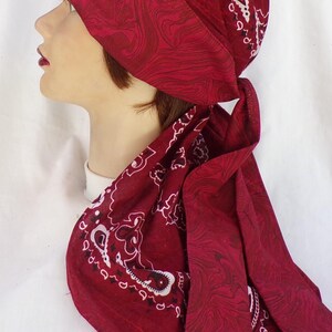 May include: A burgundy head covering with a paisley pattern and a swirling design. The head covering is tied at the back and drapes down the neck. The fabric has a white paisley design.