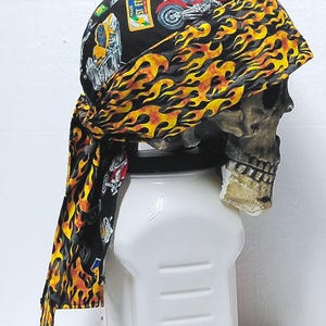 May include: A black bandana featuring a vibrant yellow and orange flame pattern along the border and a graphic print of motorcycles and rectangular patches on the crown. The fabric headwear is tied at the back with two long, trailing fabric tails.