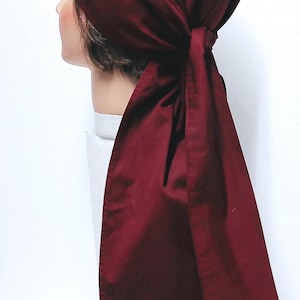 May include: A burgundy head wrap with a long, flowing tail. The head covering is made of a smooth fabric and is tied at the back. The head wrap is displayed on a mannequin head against a white background.