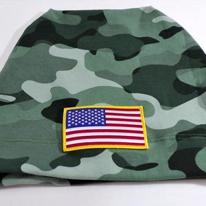 May include: A green camouflage beanie with an American flag patch. The hat features a pattern of dark and light green shapes. The flag patch is rectangular with a yellow border, displaying the stars and stripes.
