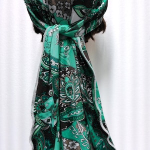 May include: A green and brown paisley patterned headscarf tied in a knot. The scarf features a repeating floral design with white and brown accents. The edges are finished with a white, decorative stitch.