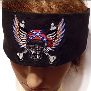 May include: Black headband featuring an embroidered skull with wings and crossbones. The skull is wearing a Confederate flag bandana. The wings are in shades of brown, blue, and white. The headband is a wide, stretchy band.