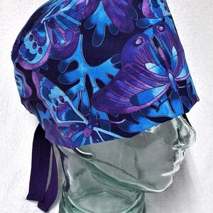 May include: A blue and purple patterned surgical scrub cap featuring a butterfly design. The cap has a dark blue background with large, stylized butterflies in shades of blue and purple. It includes a matching purple tie at the back for adjustment.