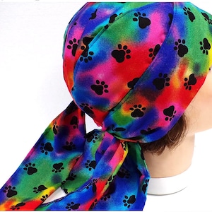 May include: A colorful head wrap featuring a tie-dye pattern in shades of red, pink, blue, green, and yellow. The fabric is adorned with black paw prints. The head wrap is tied at the back.