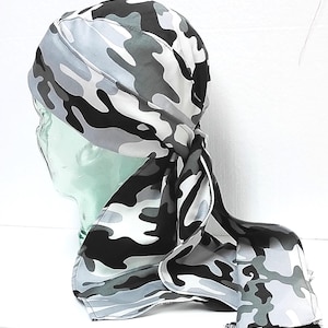 May include: A gray, black, and white camouflage pattern head wrap. The head covering is tied at the back, with long, flowing tails. The fabric has a repeating pattern of irregular shapes in shades of gray, black, and white.