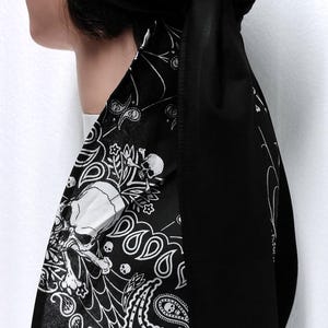 May include: Black skull bandana head wrap with a paisley design. The head wrap features a black base with white skull and paisley patterns. The head wrap is tied at the back.