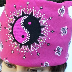 May include: A pink bandana featuring a black and pink yin-yang symbol, embellished with sparkling rhinestones. The design includes a white floral pattern around the yin-yang and smaller decorative elements.