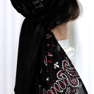 May include: Black head covering with a red and white paisley pattern. The head wrap is tied at the back, with long fabric tails hanging down. The paisley design is a classic, decorative motif.